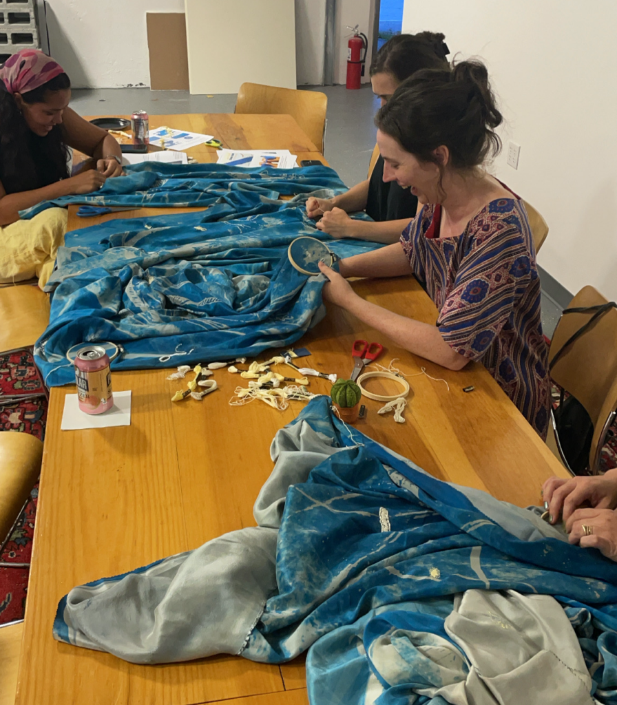 Photograph of three women and one set of hands at a long wooden table with blue and light gray swaths of fabric laid out over the table. Each women is using embroidery thread to repair parts of the fabric.