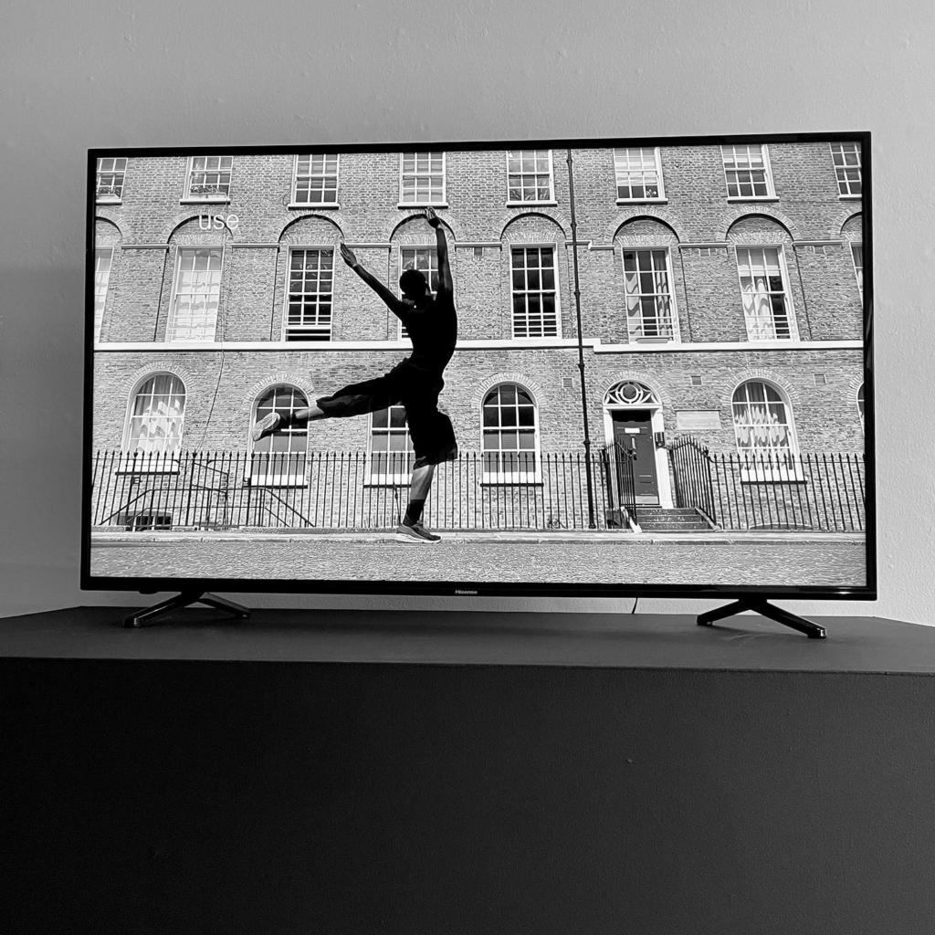 Photograph of a television screen with a black and white image of a dancer in front of a brick building.