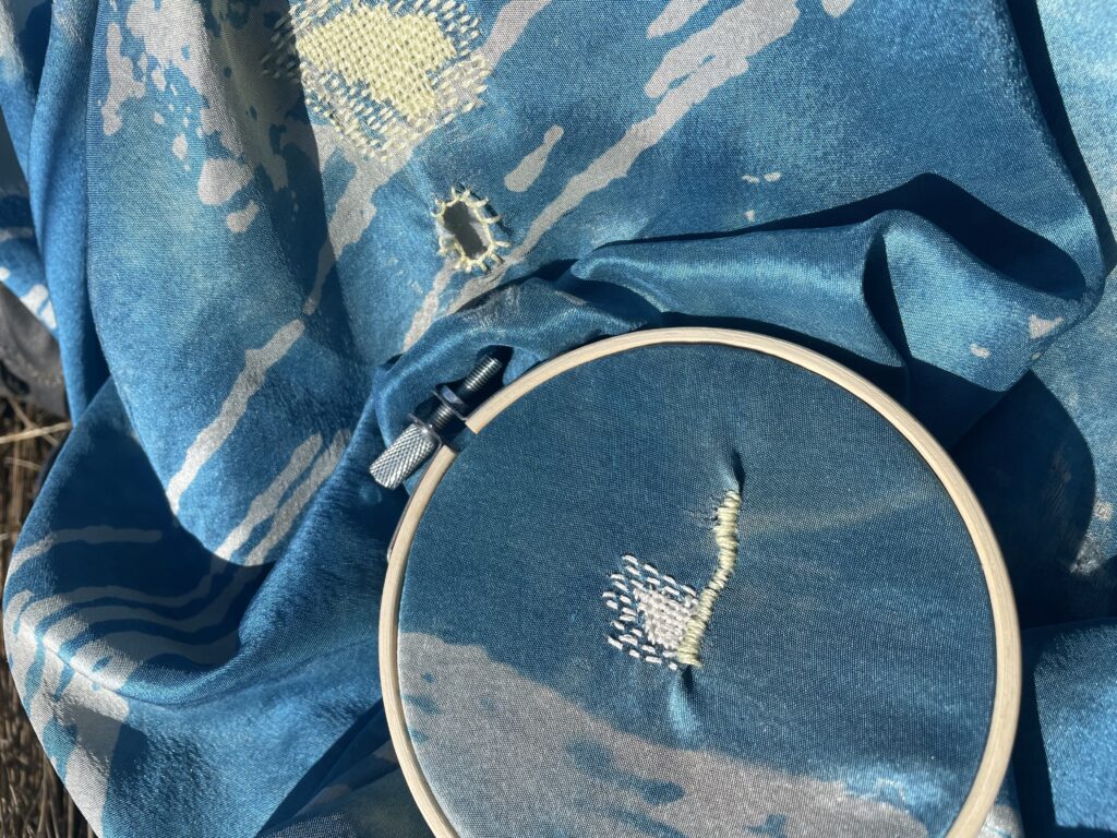Image of blue fabric with light gray pattern draped out with the lower right portion in an embroidery hoop. There are three areas that have been mended with yellow or white thread.