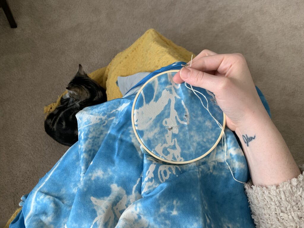 Image of blue and light gray fabric in an embroidery hoop on someone's lap. There is hand holding the hoop and a light colored thread. A cat sits below.