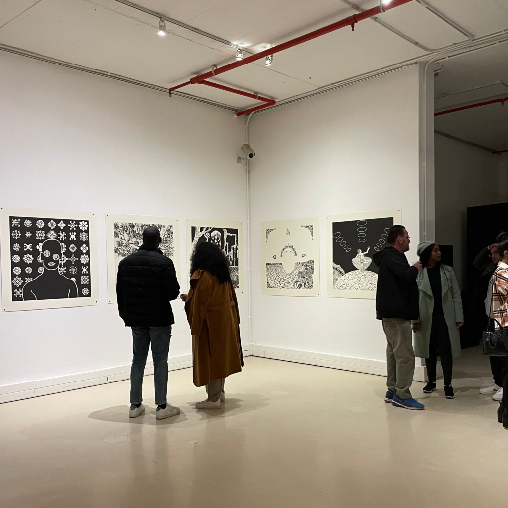 Photograph of people in a gallery viewing black and white square illustrative artwork on the wall in a gallery setting.