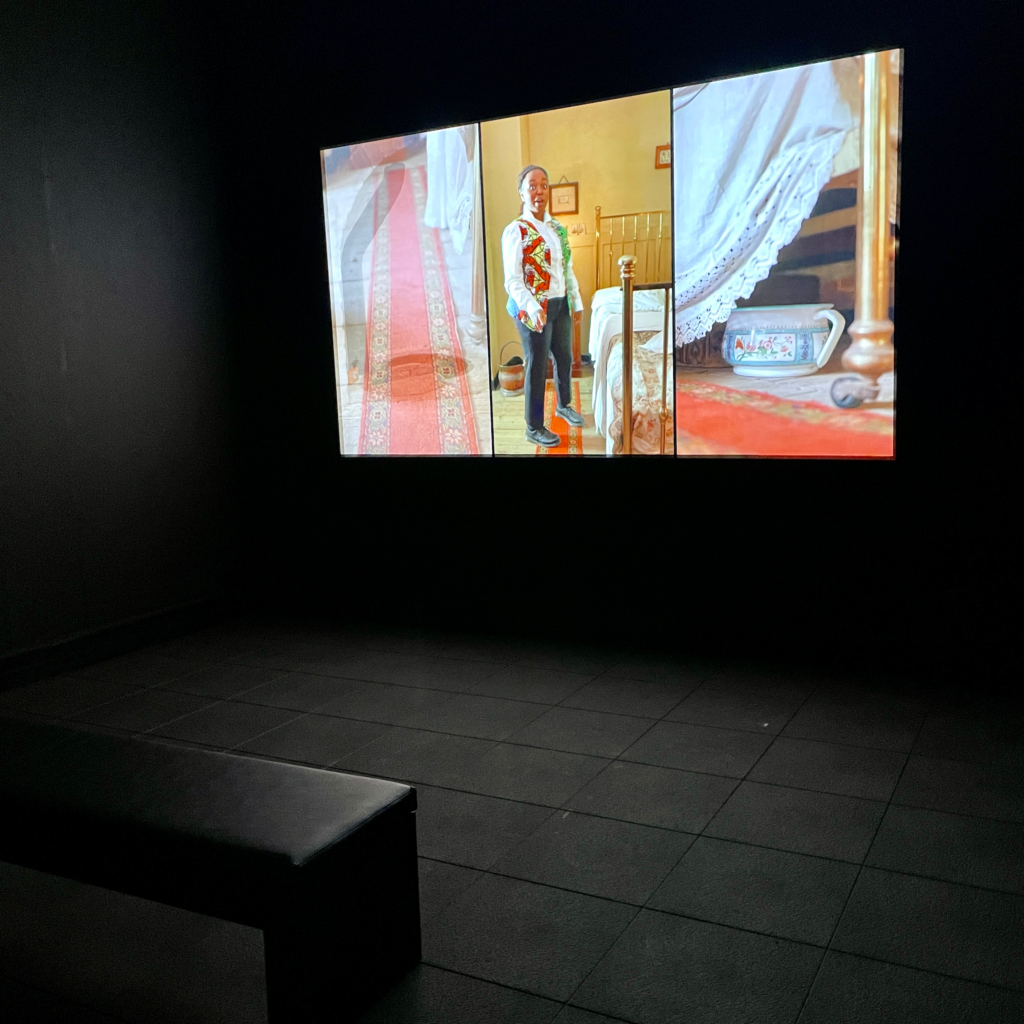 Photograph of black room with bench and a split screen video of a woman and parts of a room being projected onto the wall.