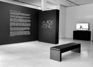 A black and white photograph of a gallery setting with an L shaped black wall with white text on the left and Black Sonic in a stylized font written in white on the right. The the right there is a black stand with a television screen depicting someone standing with legs wide in an parking lot. Facing the L shaped wall is a black bench.