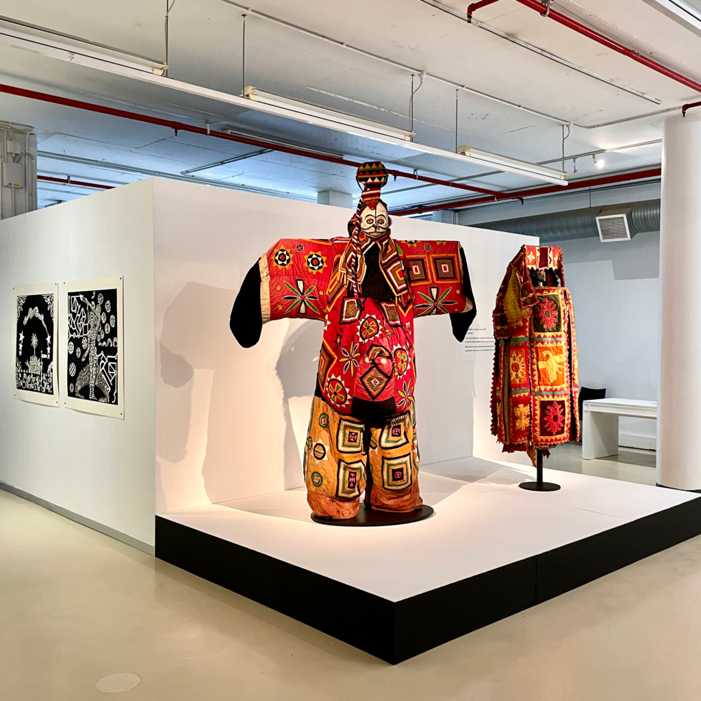 Photograph of gallery setting with two stands featuring hand made attire with geometric patterns and a palette of black, red, yellow and white. The stand on the left has a white mask with a head piece on top. On the wall around the corner to the left there are square black and white illustrative prints.