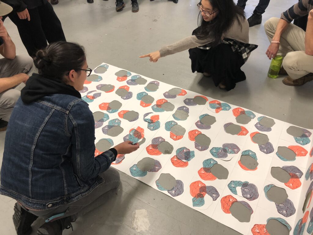 Photograph of a paper puzzle with coral, teal and purple printed amorphous shapes is laid across the floor with two crouching people on either side. The person on the top is pointing and the person at the bottom is holding a puzzle piece. Others can be seen standing or crouching outside the frame of the image.