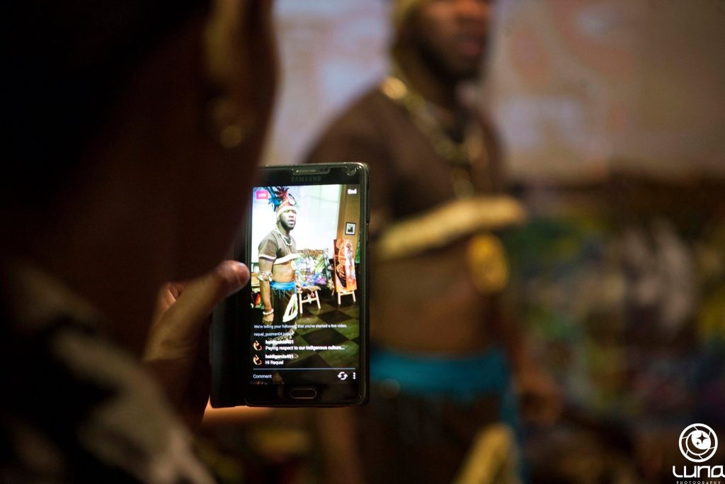 Photograph of person of color holding a phone to record a video for social media of a man of color in a headdress.