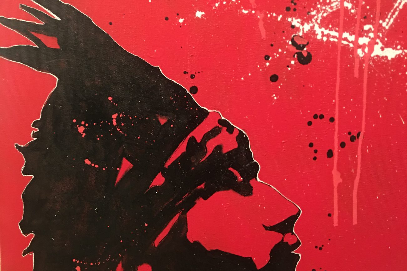 Red artwork depicting a native man from the chin up in a headdress painted in black with black, white and red splatters.