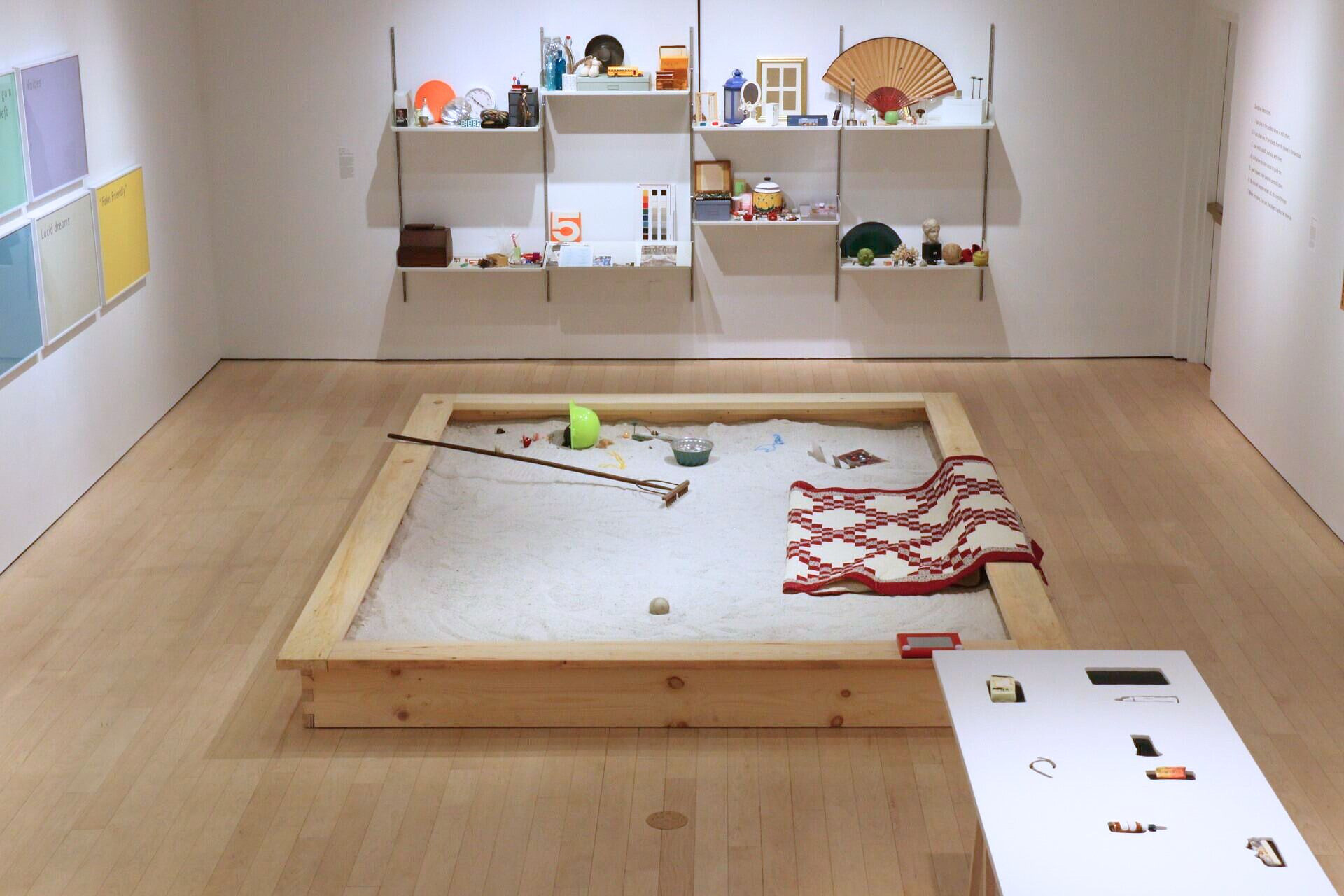Art exhibition featuring various everyday objects sitting on shelves, a white table, and more objects in a brown sandbox in the center of the room.