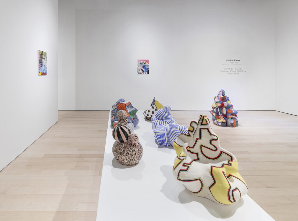 Art exhibition featuring five ceramic sculptures, consisting of various patterns and shapes, on a white pedestal. Abstract artworks hanging on walls and a multicolored irregular shape sitting on the floor.