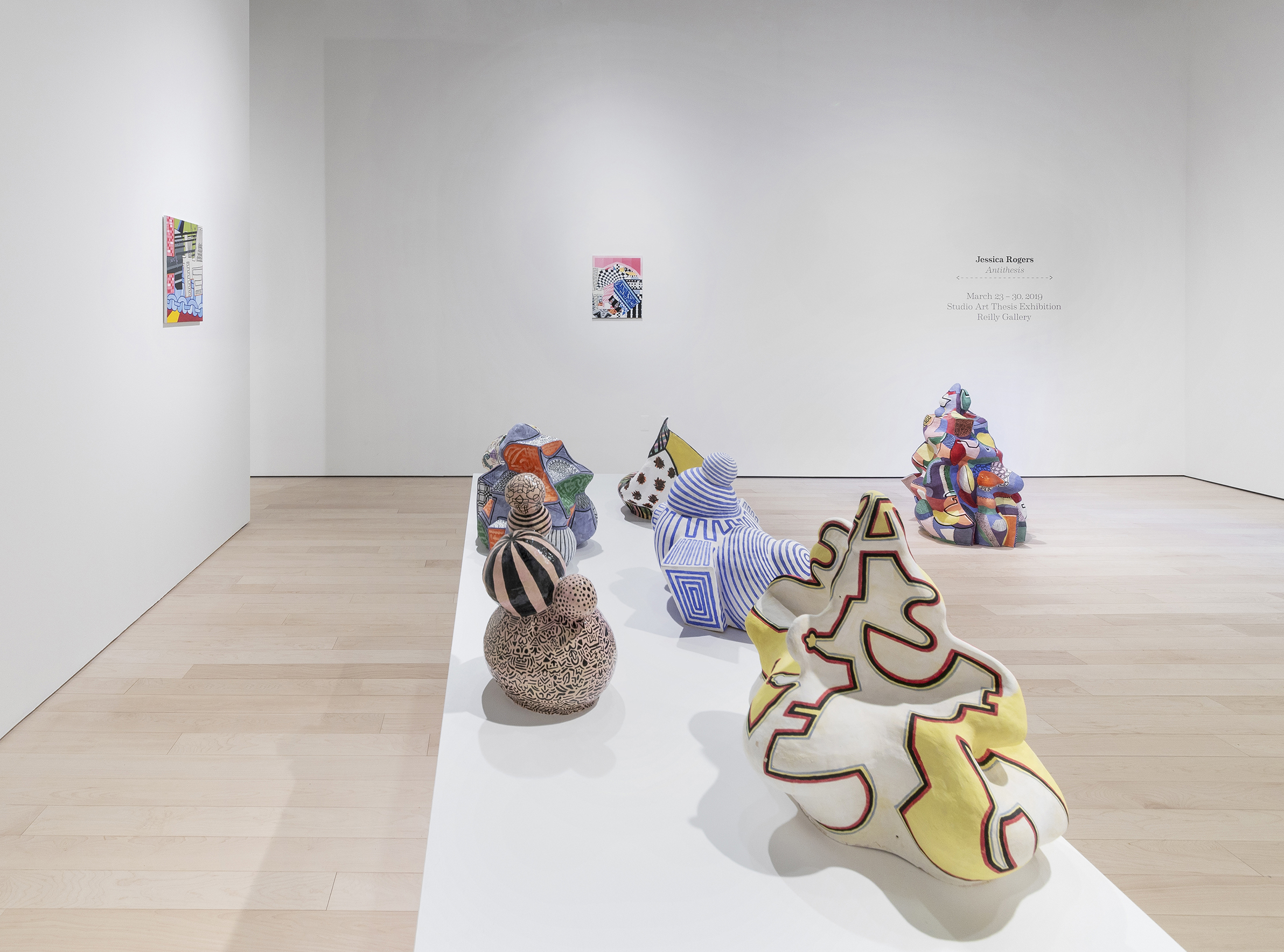 Art exhibition featuring five ceramic sculptures, consisting of various patterns and shapes, on a white pedestal. Abstract artworks hanging on walls and a multicolored irregular shape sitting on the floor.