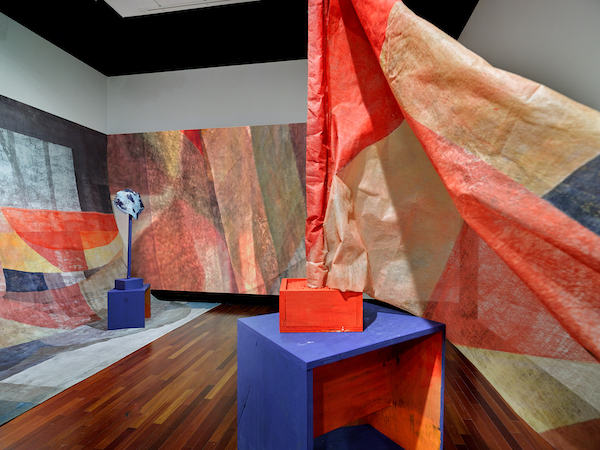 Installation view of a bright blue and orange table-like structure with a large orange abstract flag cascading from the top. Artwork with similar coloring covers the back walls.