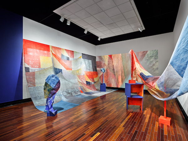 Installation view of large expanses of fabric filled with brightly colored shapes covering two walls. To the right, orange and blue structures are holding up more fabric with similar colors.