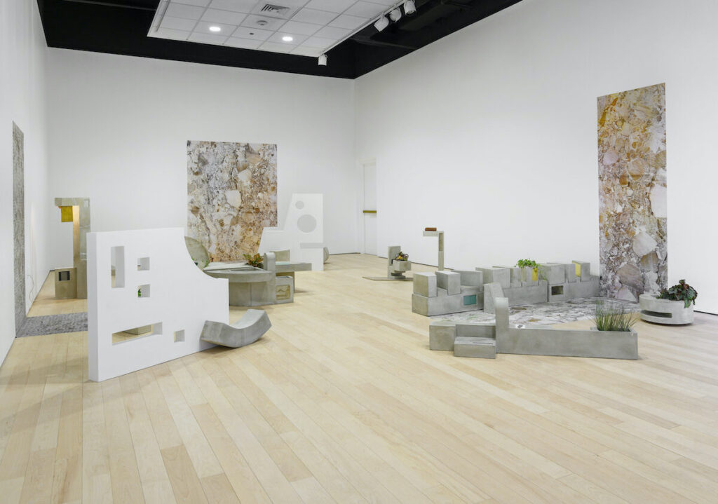 Installation view of abstract sculptures of large white geometric shapes and smaller grey blocks. The right and back wall each have a rectangular artwork of what looks like colorful rocks.