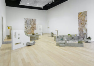 Installation view of abstract sculptures of large white geometric shapes and smaller grey blocks. The right and back wall each have a rectangular artwork of what looks like colorful rocks.