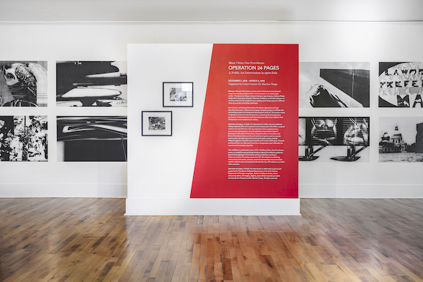 Art exhibition featuring a short white wall with half of it red and two black-and-white frames hanging. The taller wall in the back holds eight black-and-white prints not framed.