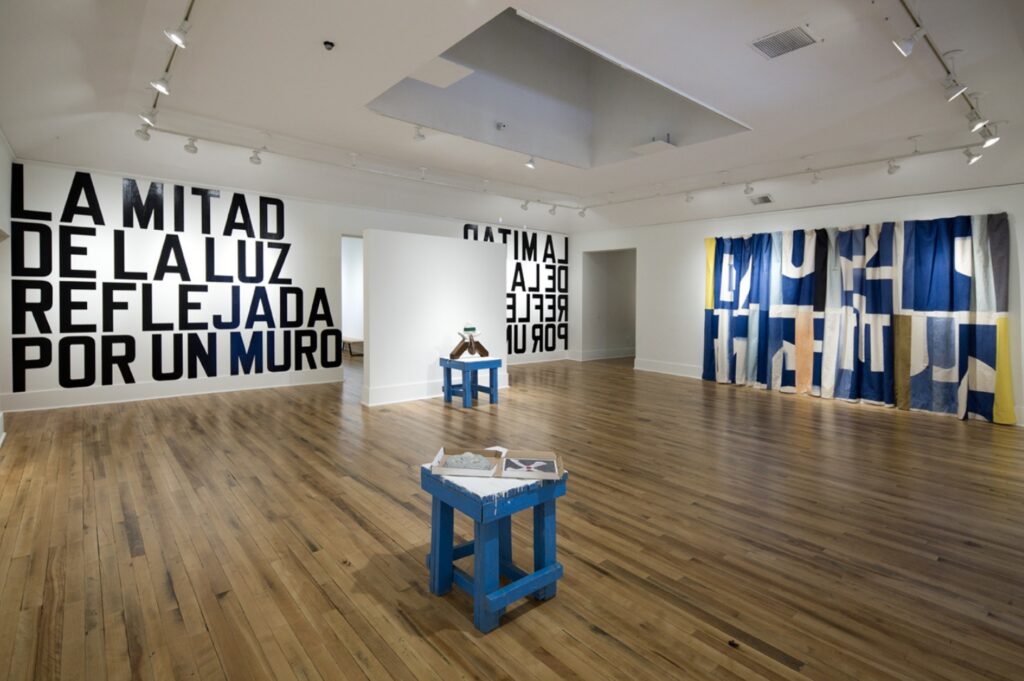 Installation view with two blue stools, one in the foreground, one in the background. The back wall reads LA MITAD DE LA LUZ REFLEJADA POR UN MURO. The right wall is covered with a large abstract patterned fabric.