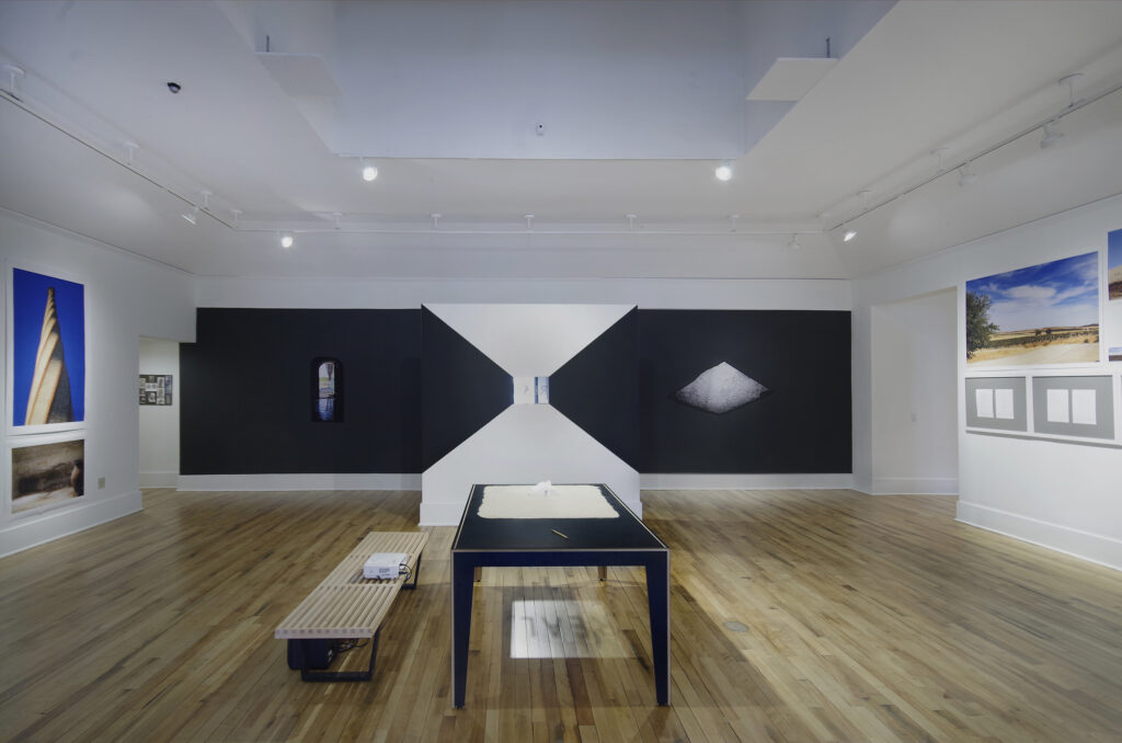 Art exhibition featuring a black-and-white wall with three small images in the distance. Closer to the camera, a black table holds yellow sand and a white object. Beneath the table is a reflection of the word REAL.
