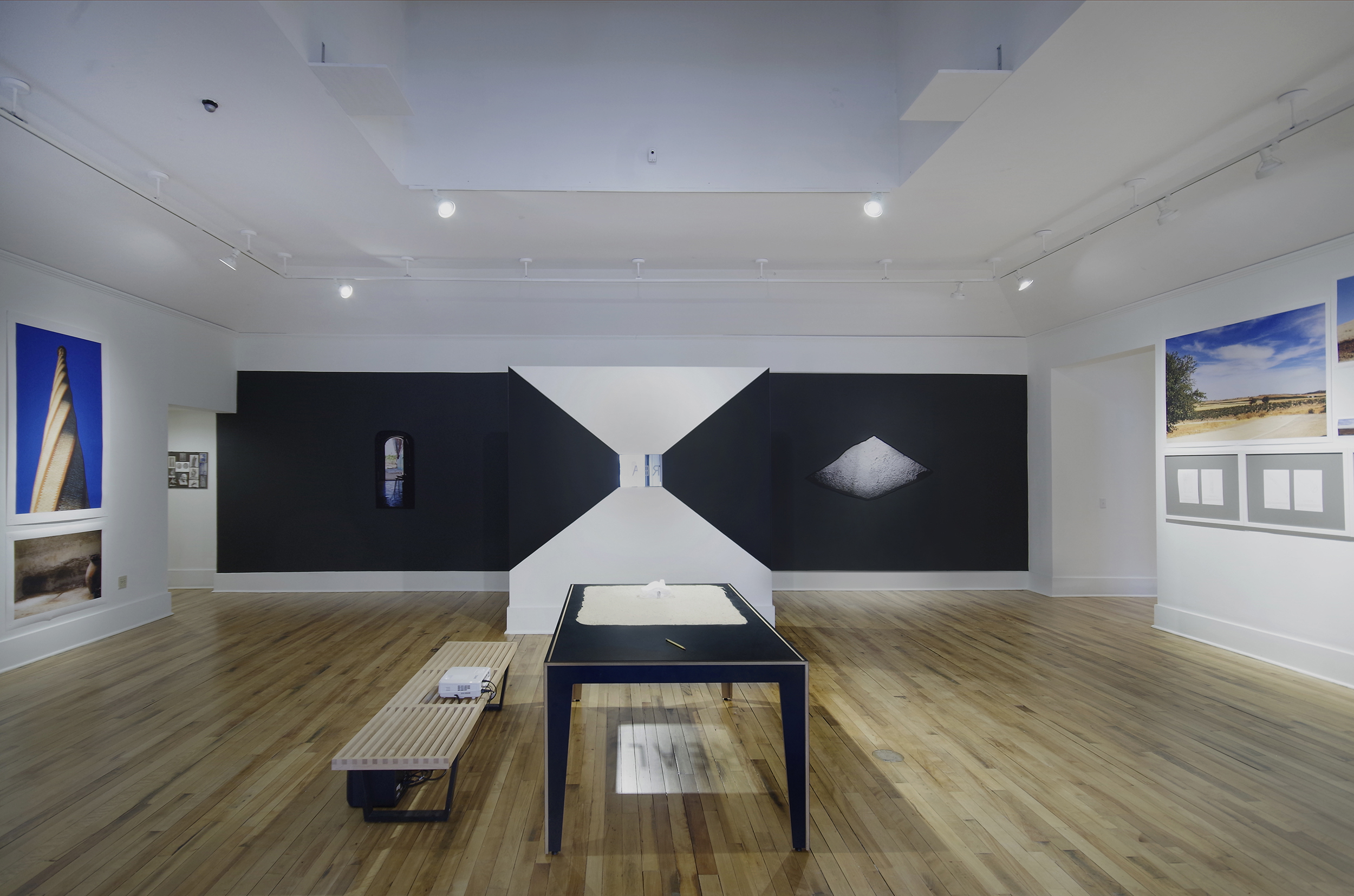 Art exhibition featuring a black-and-white wall with three small images in the distance. Closer to the camera, a black table holds yellow sand and a white object. Beneath the table is a reflection of the word REAL.