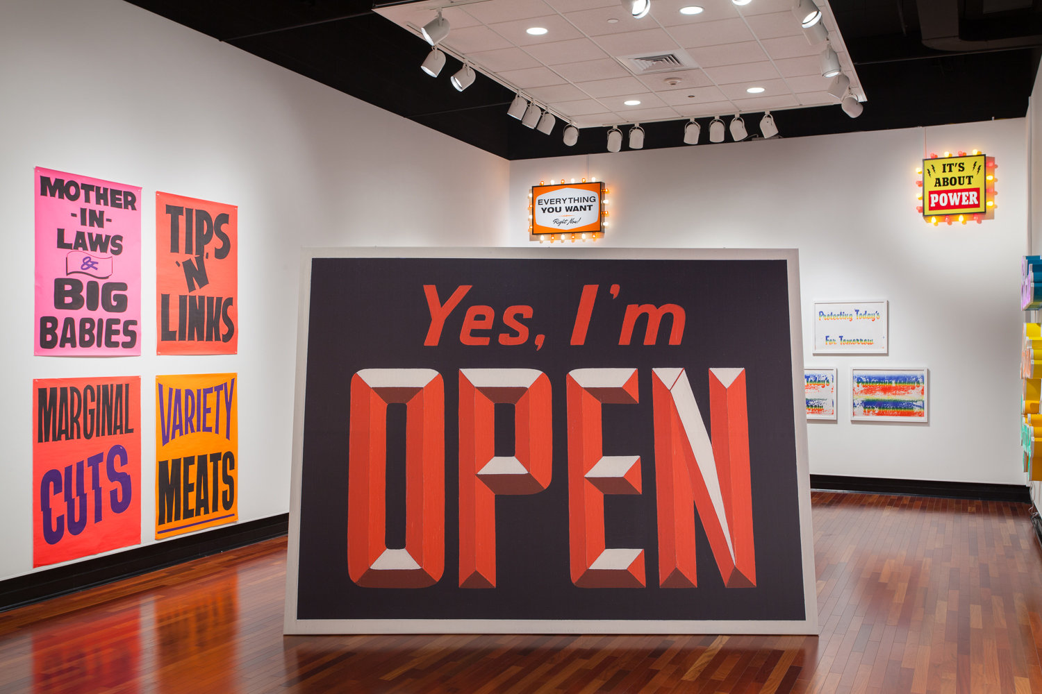 A large sign reading YES, I'M OPEN in red on a black background, in the middle of a gallery. The walls show more colorful signs.