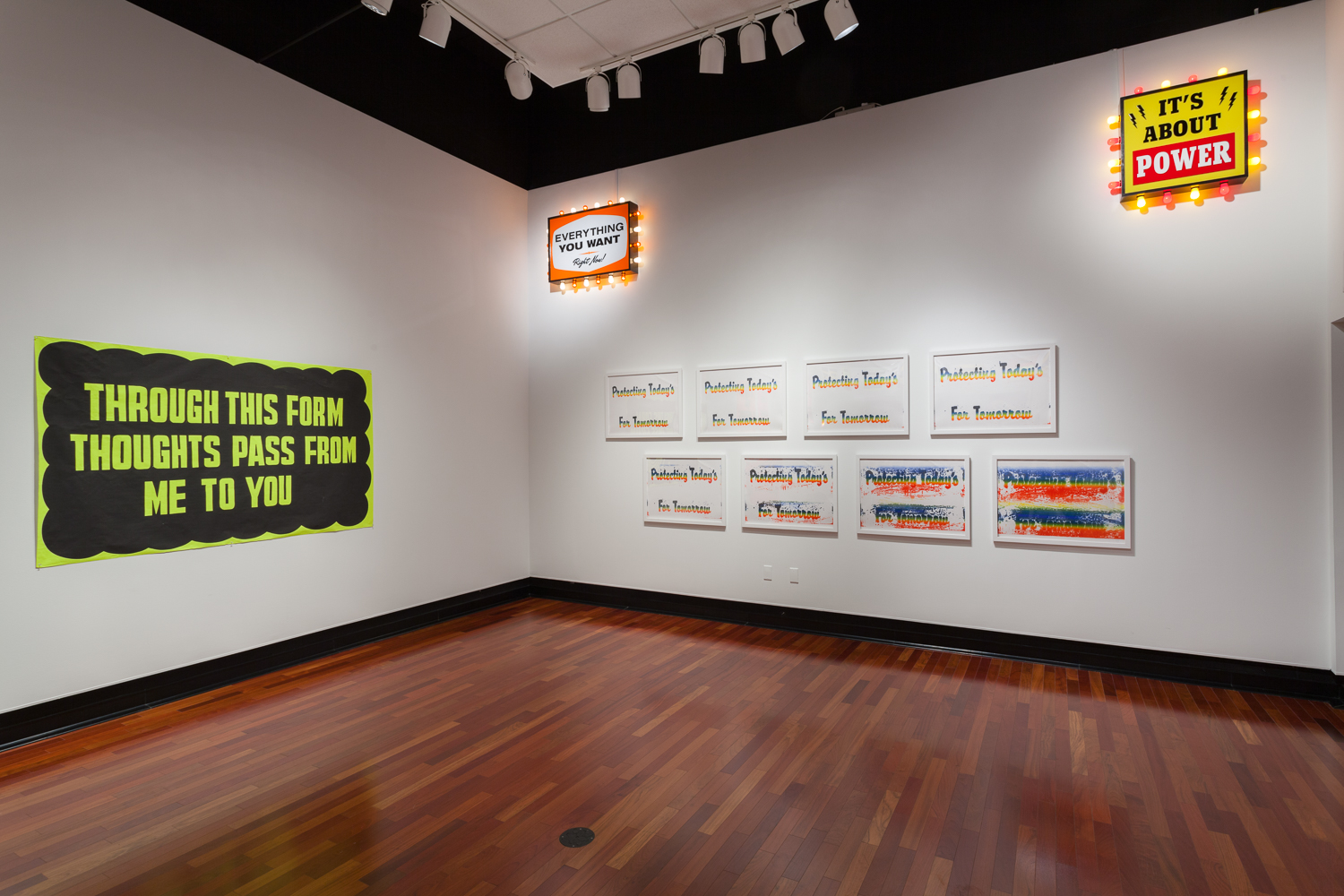 Two white walls - the left with a black sign with neon yellow words, THROUGH THIS FORM THOUGHTS PASS FROM ME TO YOU. The right wall has two rows of four signs, and lighted signs are in the top corners.