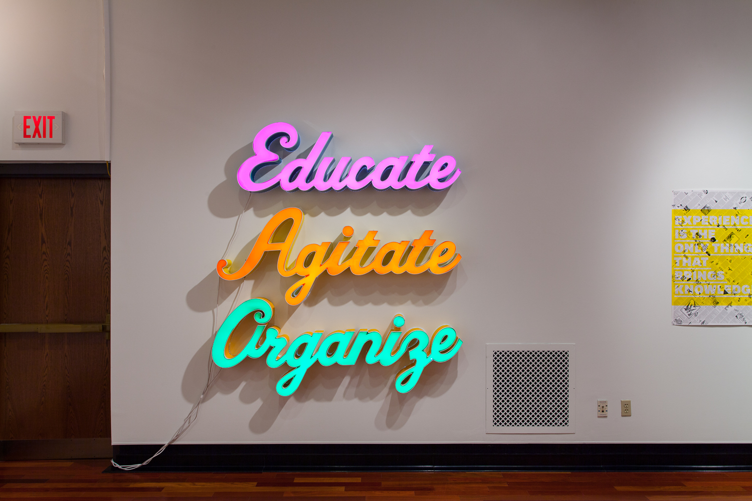 A lit neon sign of three large words in cursive stacked on top of each other on a white wall, reading EDUCATE AGITATE ORGANIZE. EDUCATE is pink, AGITATE orange, and ORGANIZE bright blue-green.