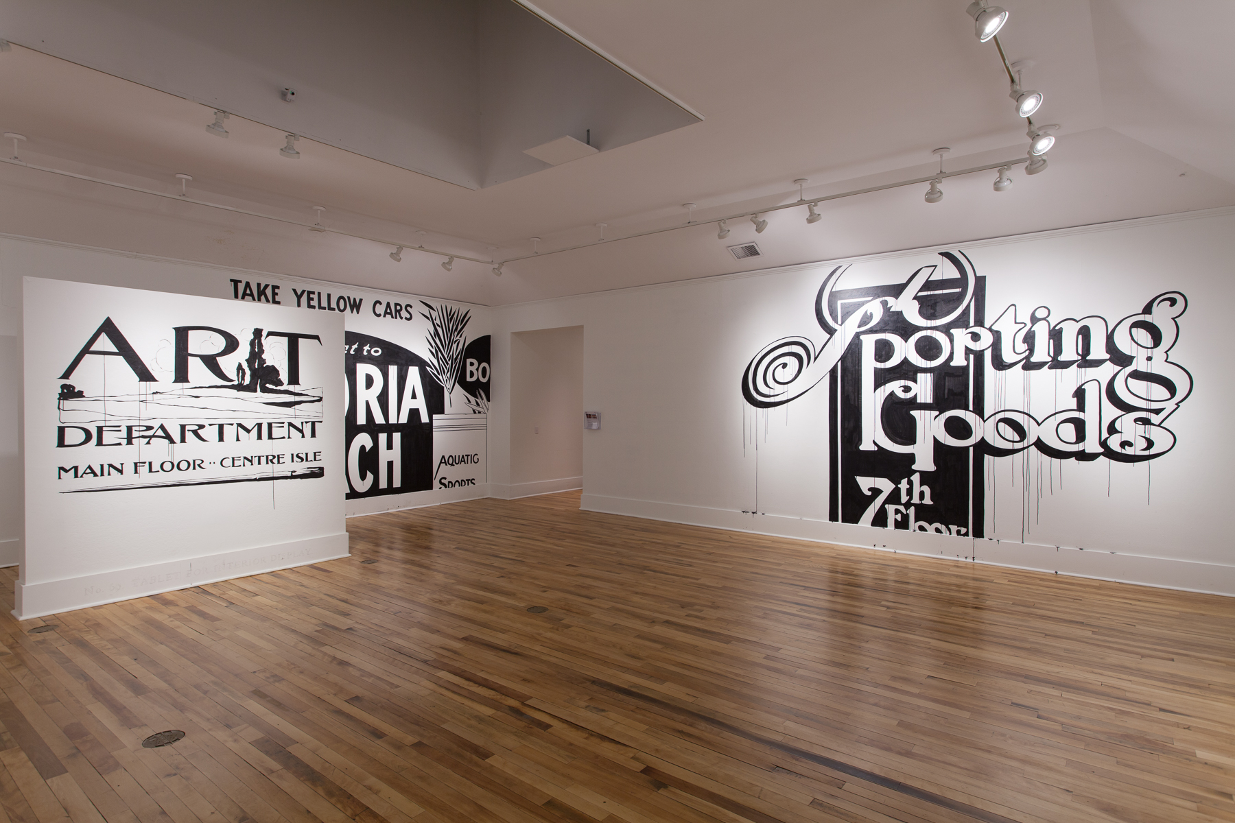 Installation view of white walls with large black and white lettering directly on them. The left reads ART DEPARTMENT MAIN FLOOR CENTRE ISLE, the right SPORTING GOODS 7TH FLOOR.