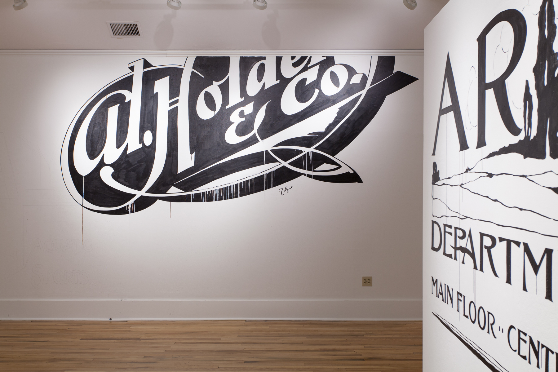 Installation view of a white wall with large black and white writing cut off at the top and surrounded by decoratve swirls. The right wall shows snippets of more writing.
