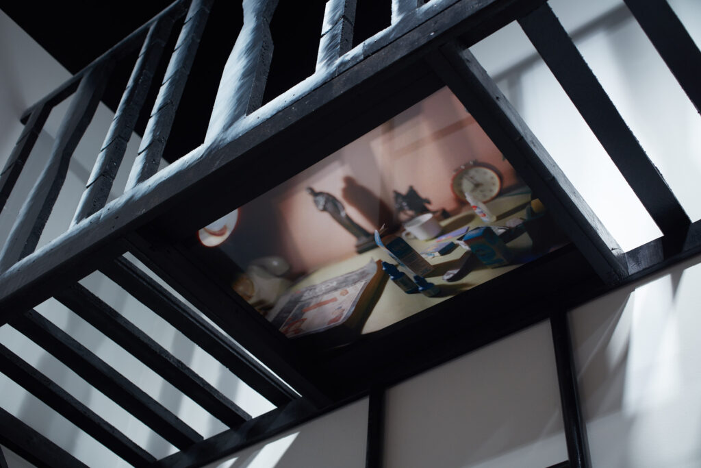 Close up of the underside of a black balcony with a railing and slatted floor. An image of everyday objects is attached to the bottom, including eyedrops and a clock.