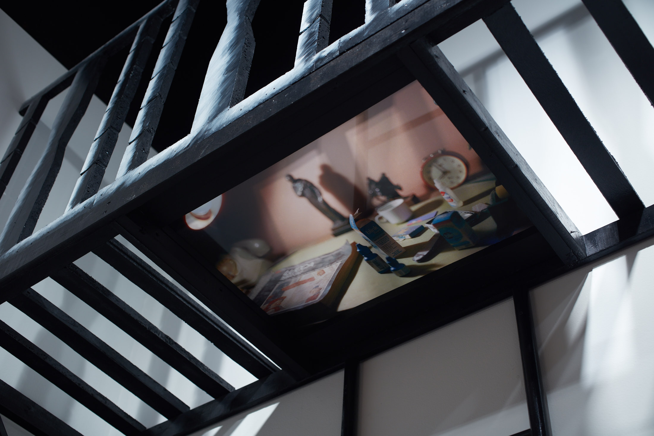 Close up of the underside of a black balcony with a railing and slatted floor. An image of everyday objects is attached to the bottom, including eyedrops and a clock.