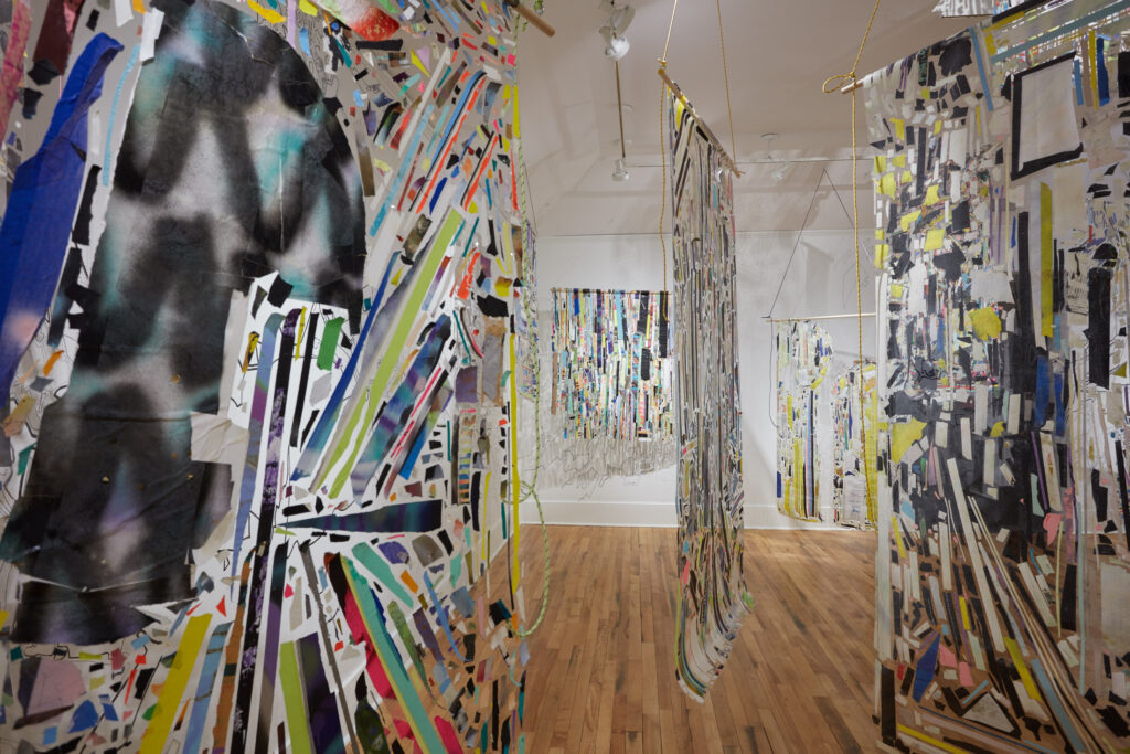 Installation view of large abstract artworks suspended from the ceiling composed of colorful strips of material. Each is attached to a wooden horizontal rod connected to rope.