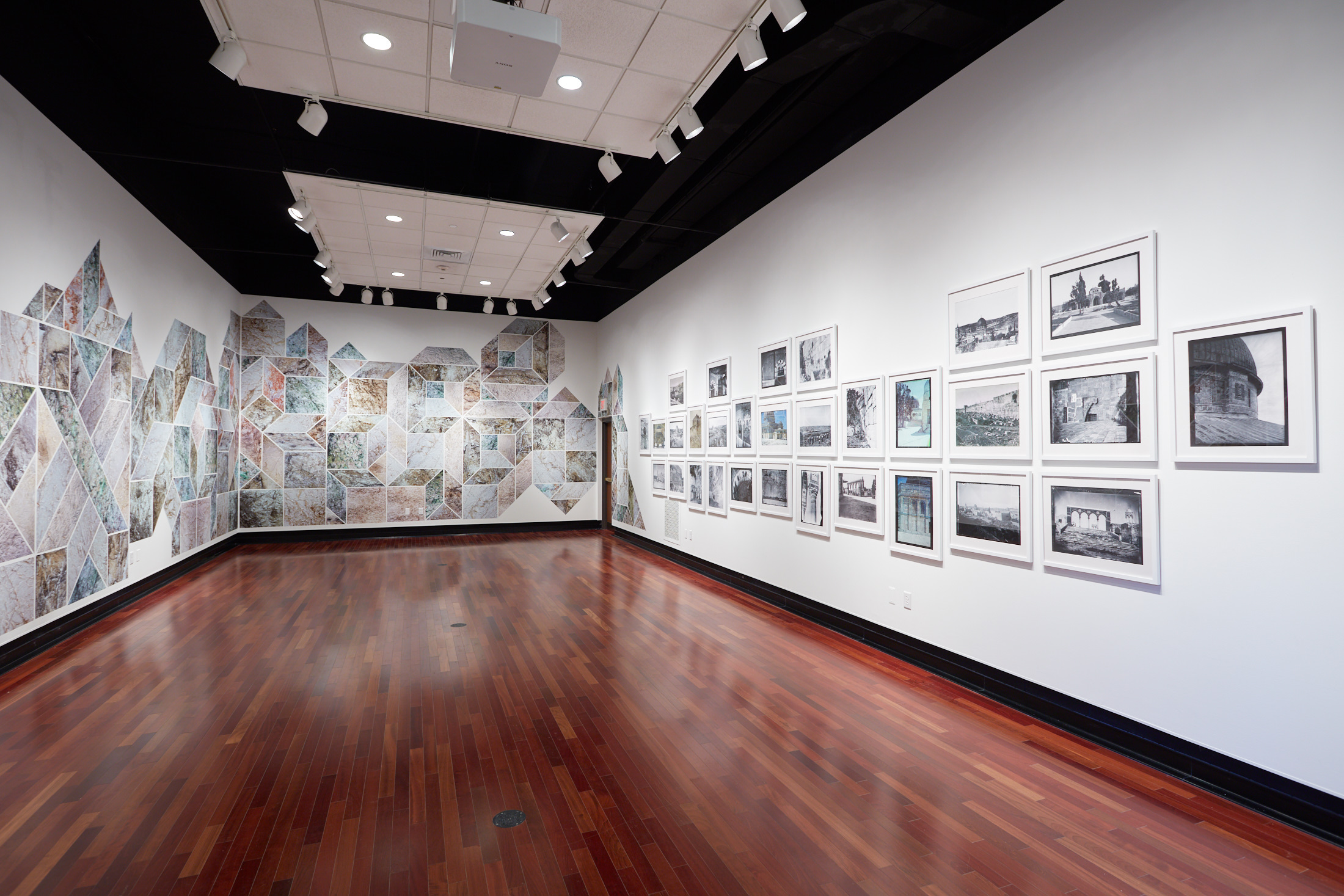 Art exhibition featuring a geometric design crossing two adjacent white walls. To the right are numerous photographs.