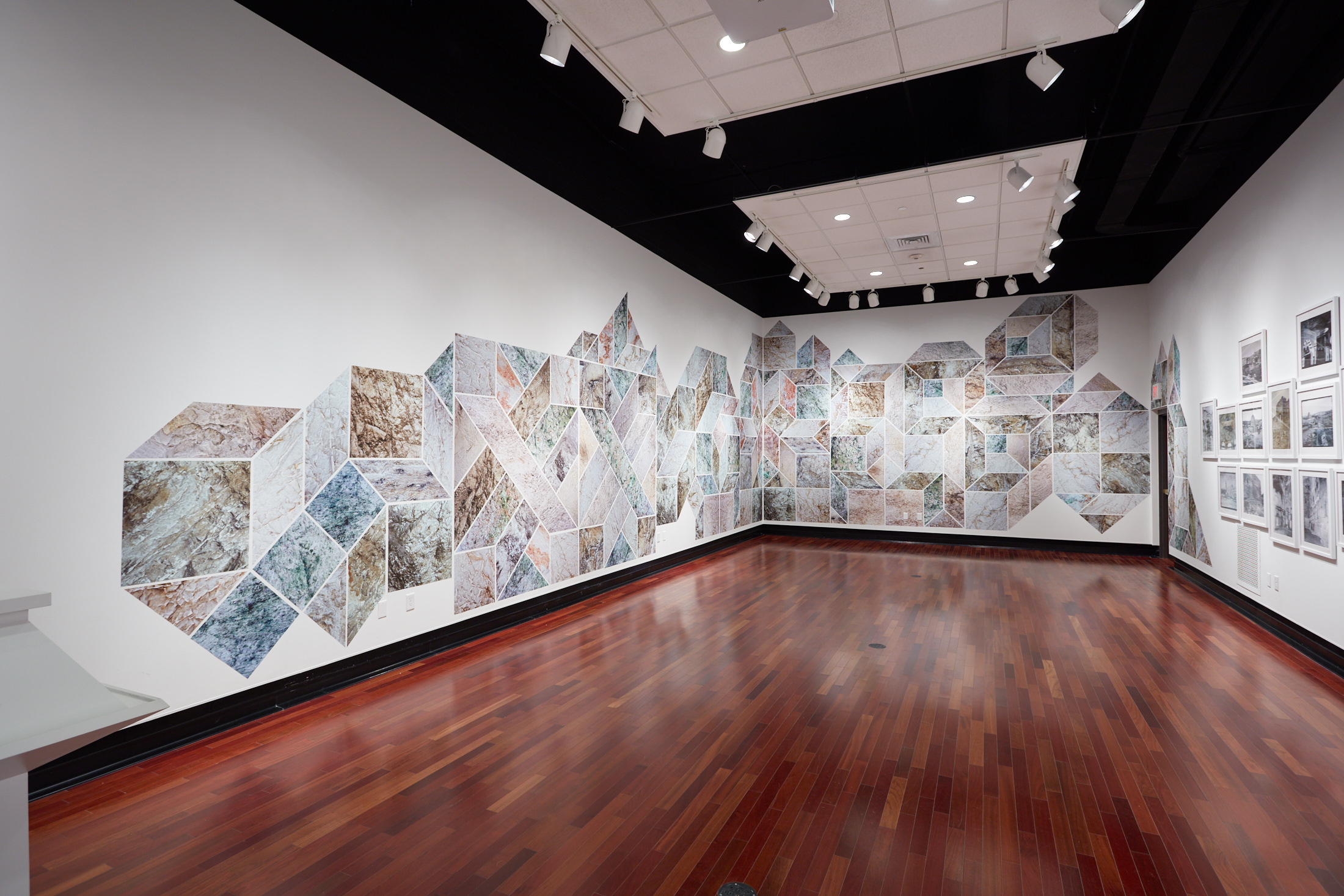 Art exhibition featuring a geometric design crossing two adjacent white walls. Off to the right are numerous photographs.