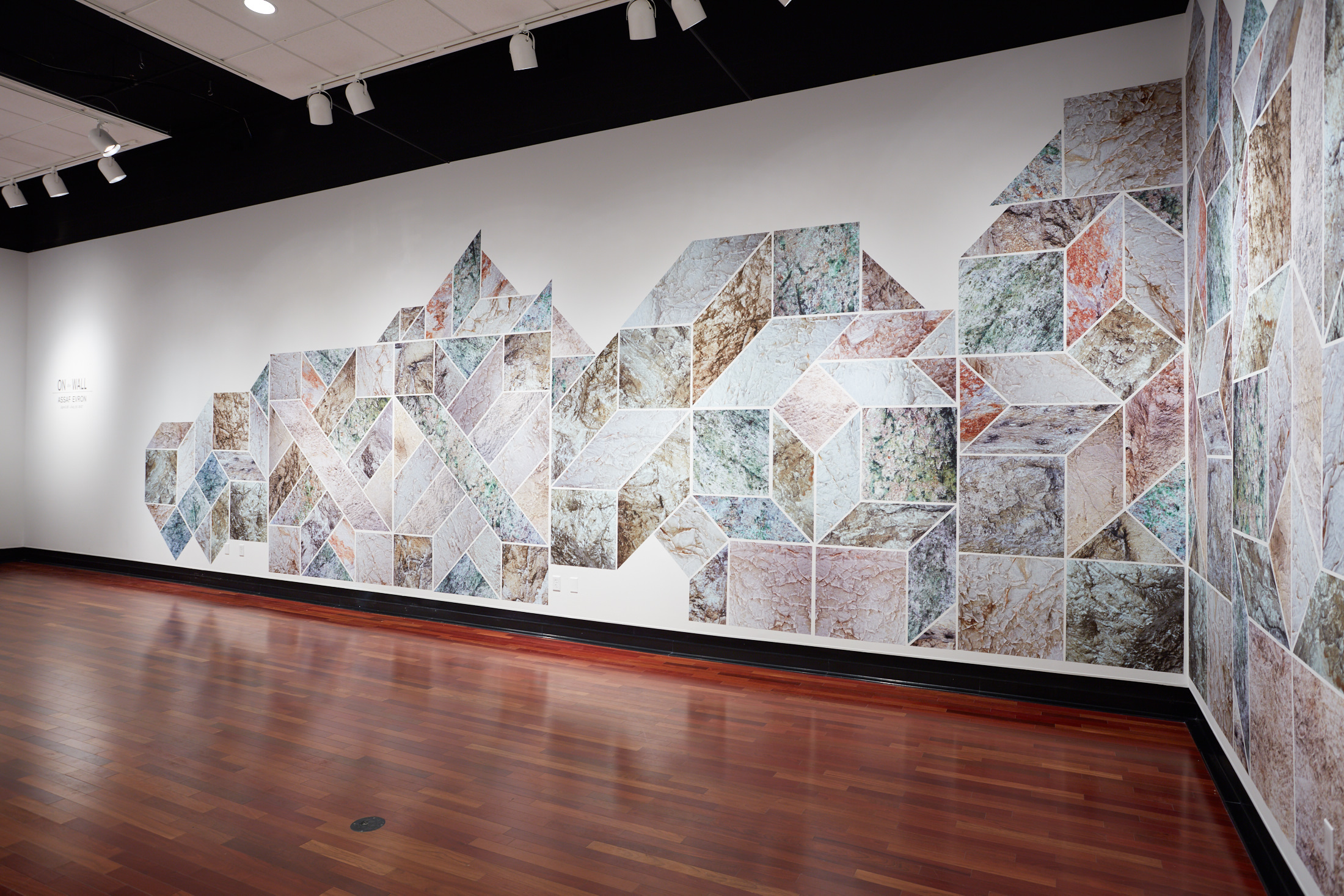 Two adjacent white walls connecting through a geometric design made up of green, orange, and beige geometric shapes, including squares, parallelograms, and diamonds.