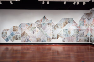Two adjacent white walls connecting through a geometric design made up of green, orange, and beige geometric shapes, including squares, parallelograms, and diamonds.