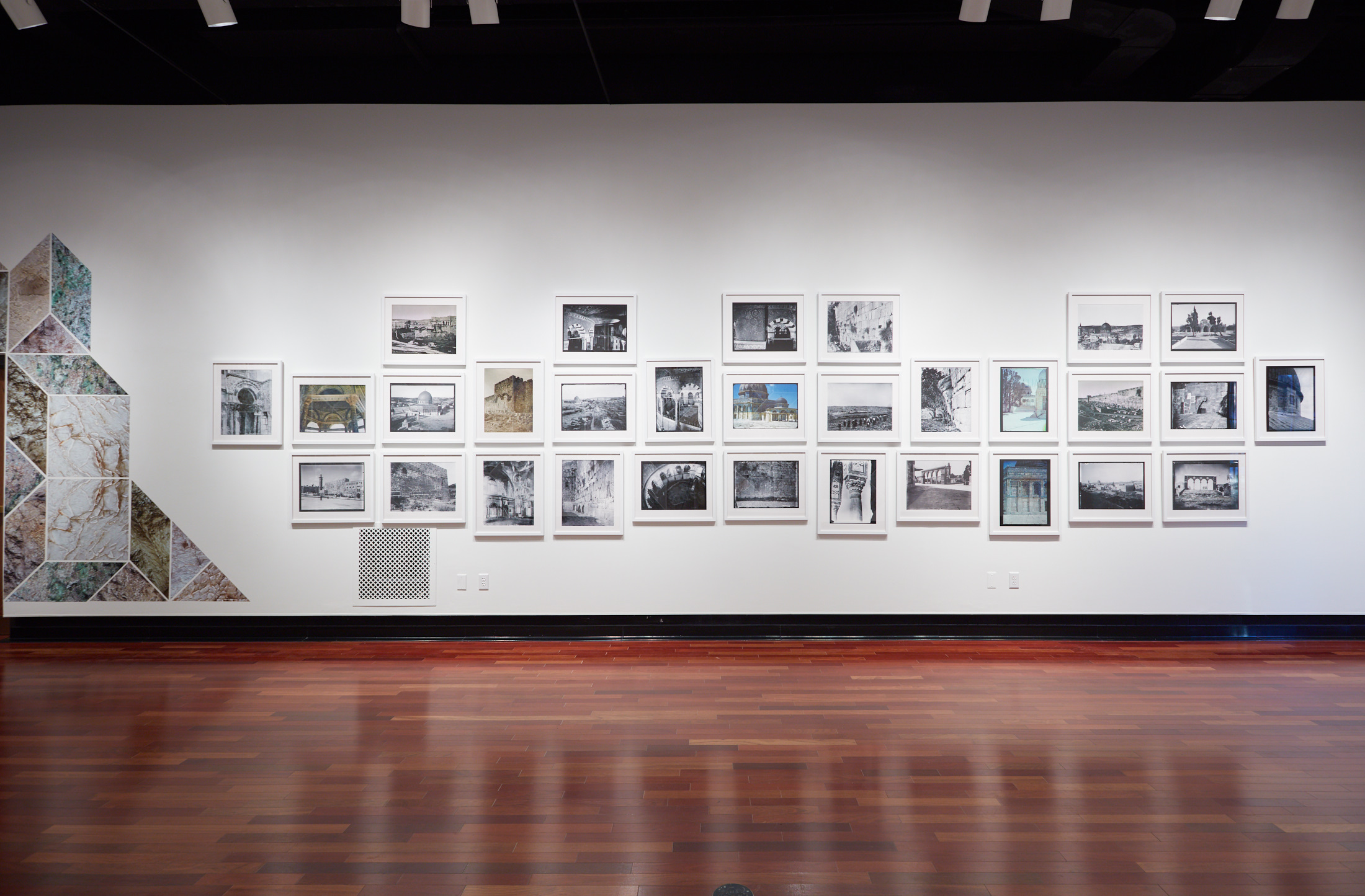 An art gallery wall featuring thirty framed photographs in a grid, mostly black-and-white with a few in color, flanked on the left by a large, triangular stone-patterned mosaic.