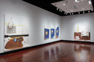 Corner featuring two white walls; the left wall displays one large and three smaller canvases, while the back wall shows a large artwork depicting construction tools.