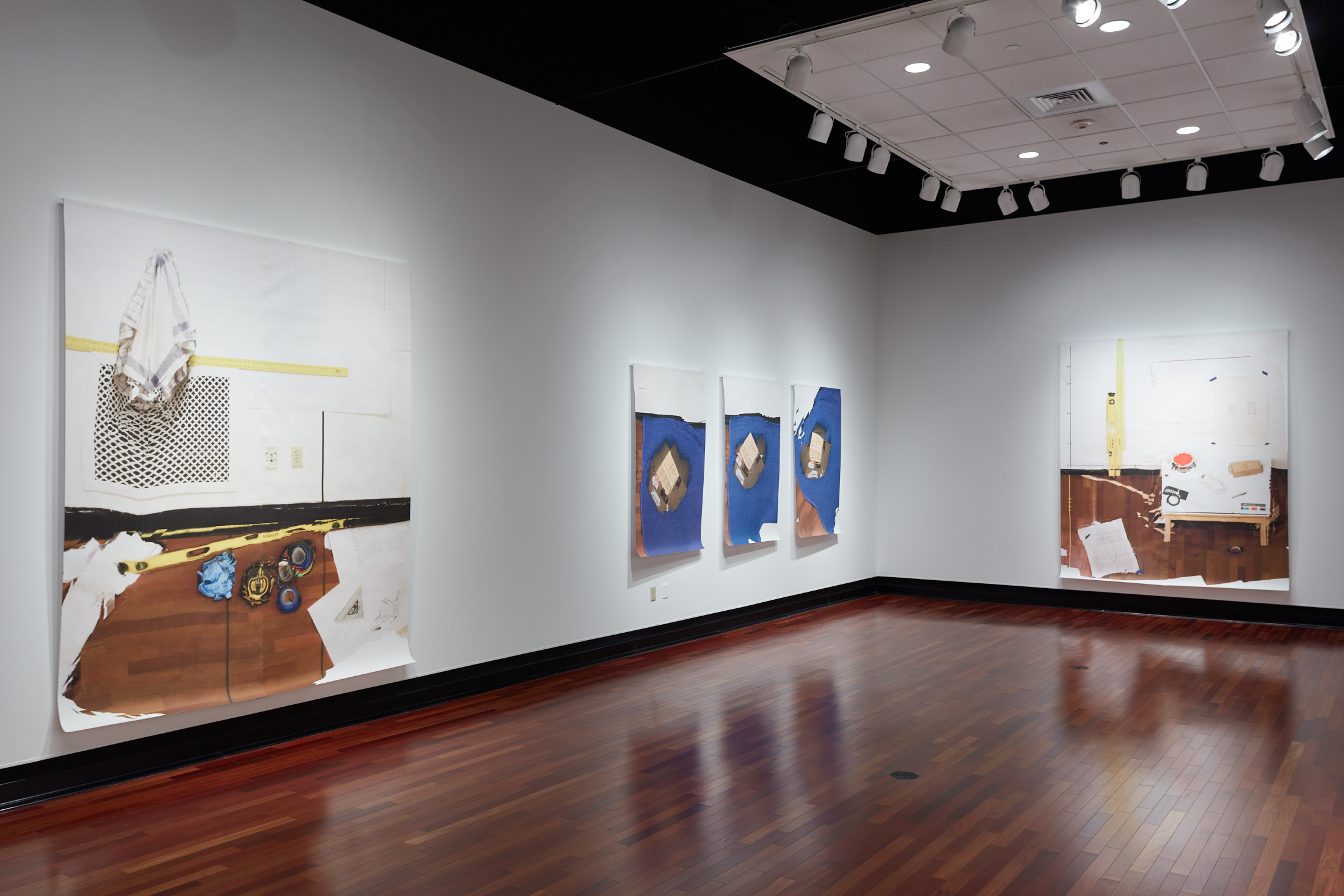 Corner featuring two white walls; the left wall displays one large and three smaller canvases, while the back wall shows a large artwork depicting construction tools.