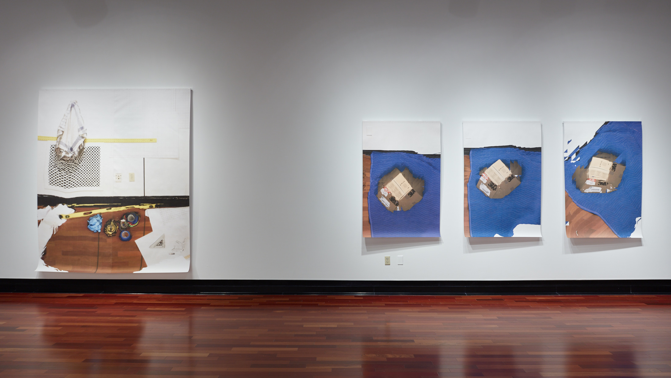 A white gallery wall with shiny wooden floors. The walls hold four canvases, one on the left showing a level, tape, and other painting and construction gear within a gallery scene. T he other three showing a box labeled "3M"with a blue blanket laying beneath it.