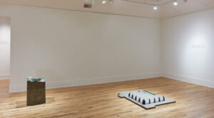A gallery with white walls and a wooden floor, with two sculptures on the ground. One is stone with smooth sides and a jagged top. The other is a smooth sltone slab with spikes.