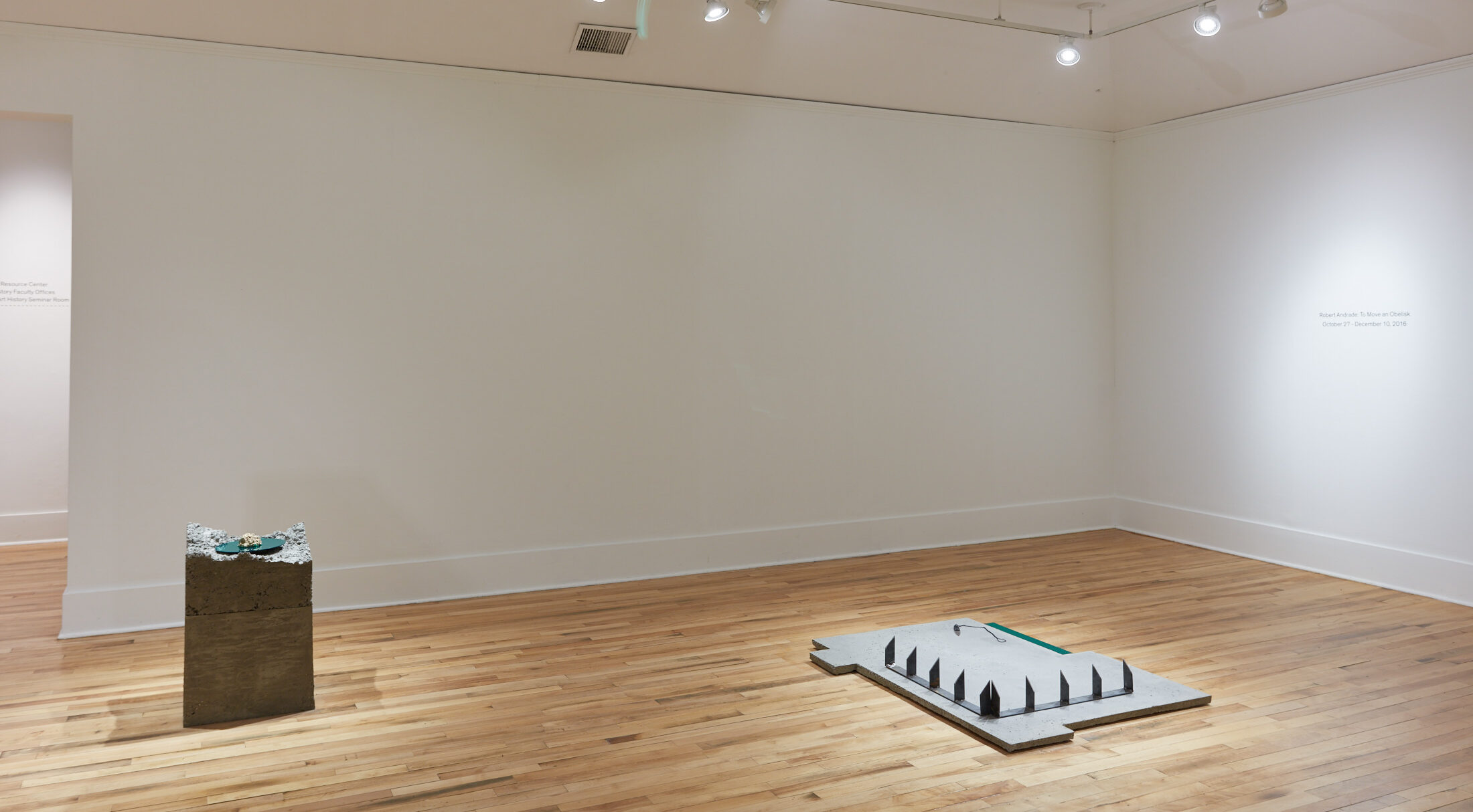 A gallery with white walls and a wooden floor, with two sculptures on the ground. One is stone with smooth sides and a jagged top. The other is a smooth sltone slab with spikes.