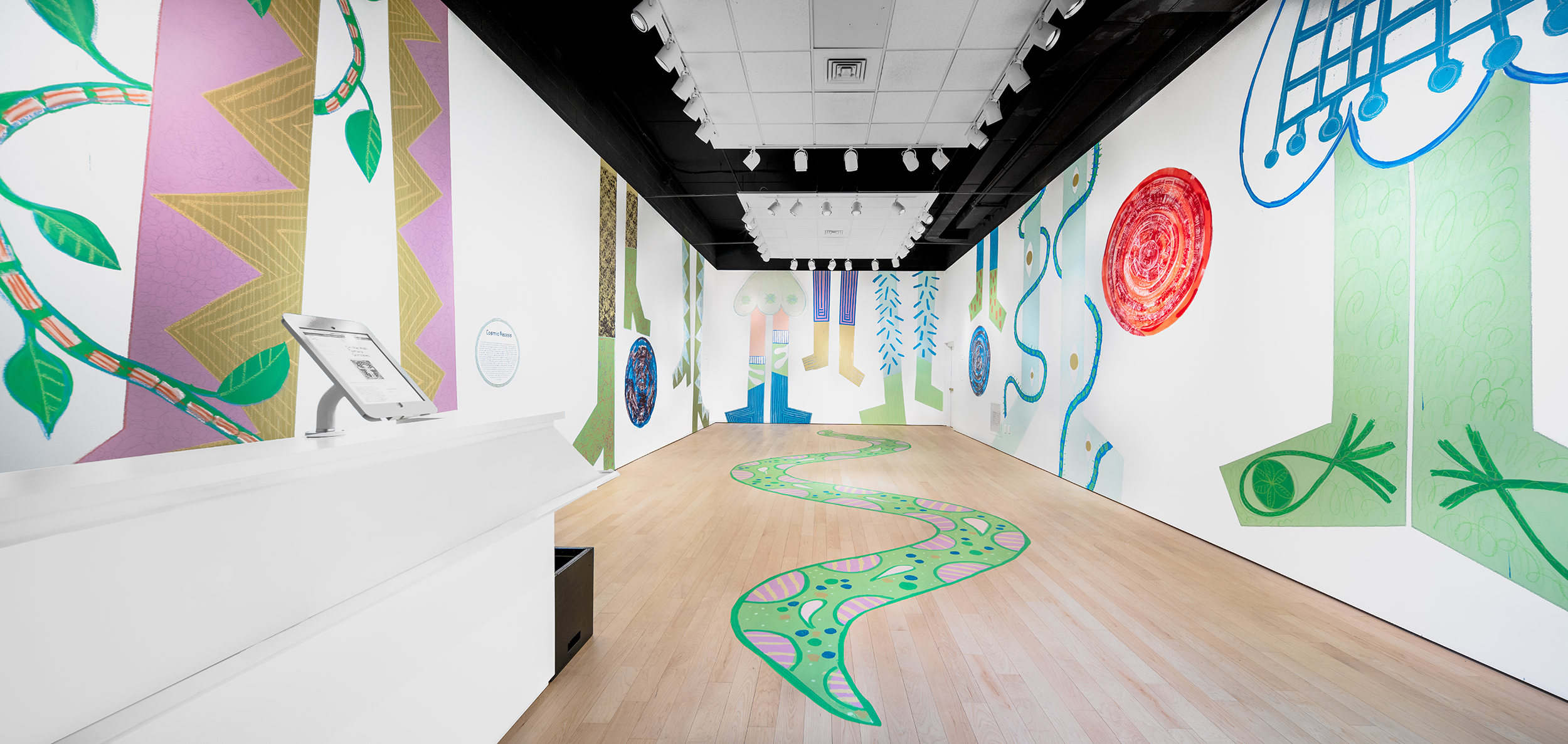 Installation view of walls painted with seven pairs of geometric legs and two large circles, one blue and one red. A green curving path is painted on the floor.