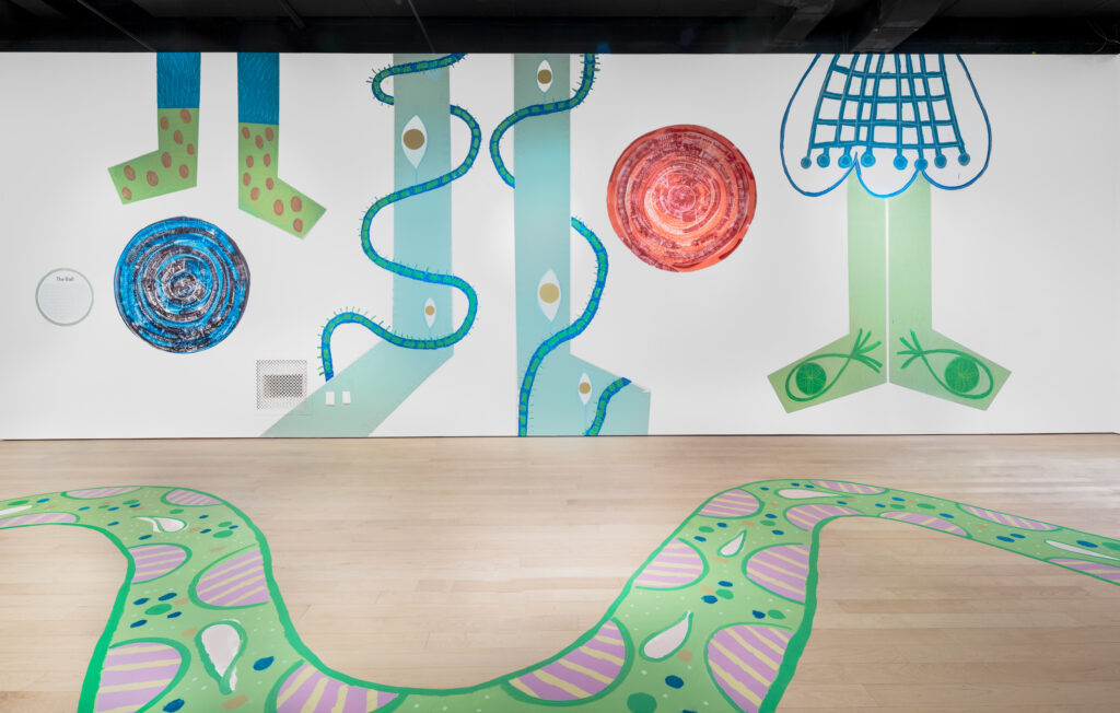 A gallery space with long cartoonish legs and feet painted on the wall in green and blue. A green, snake-like figure is painted on the gallery floor, winding up and down.