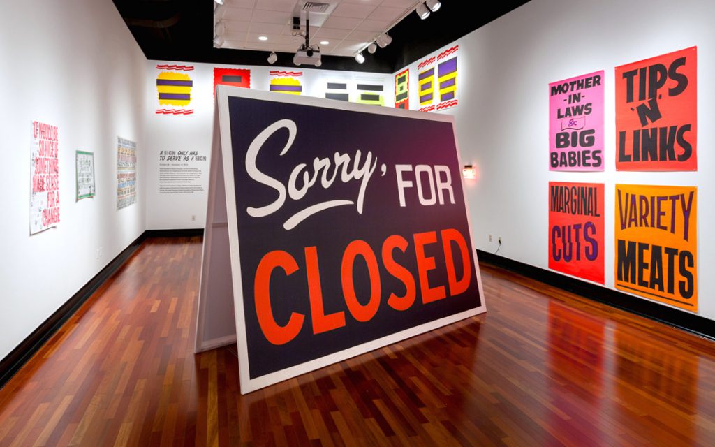 A gallery with a large tent-style sign taking up the majority of the space, with a black background. It reads SORRY, FOR CLOSED, with CLOSED in red. The walls are covered in colorful signs.