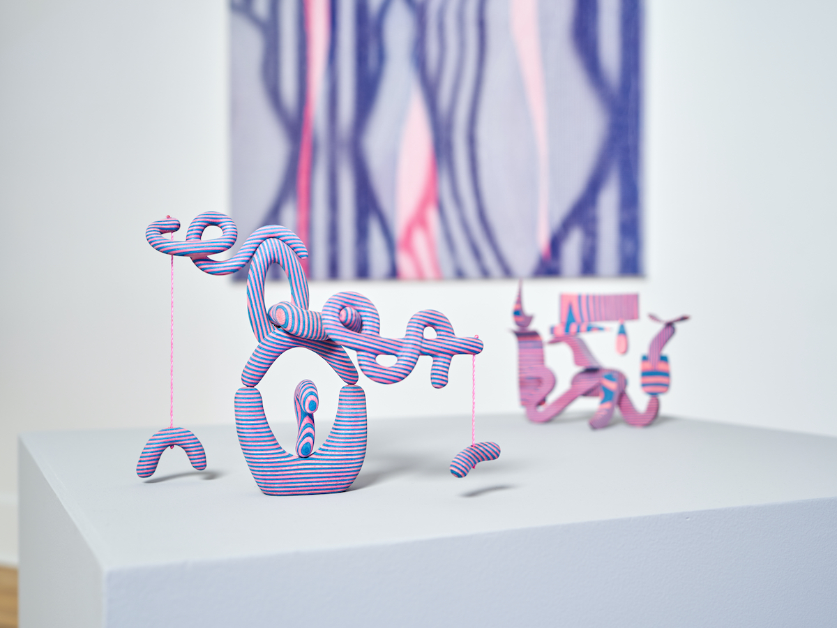 Two abstract sculptures of twisting blue and pink striped shapes - one in the foreground and one in the background. On the wall is an artwork of purple and pink curving lines.