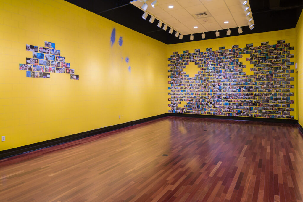 An empty gallery with yellow brick-like walls. The left wall has some photographs covering the brick, and the back wall is almost completely covered.