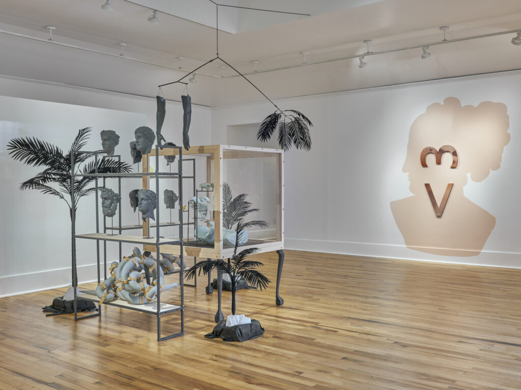 Art exhibition featuring plants and shelves of gray ceramic heads, hanging arms, and abstract objects. A large pink head silhouette on the wall is marked by a vertical heart symbol.