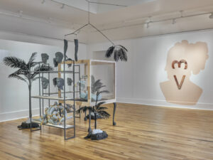 Art exhibition featuring plants and shelves of gray ceramic heads, hanging arms, and abstract objects. A large pink head silhouette on the wall is marked by a vertical heart symbol.