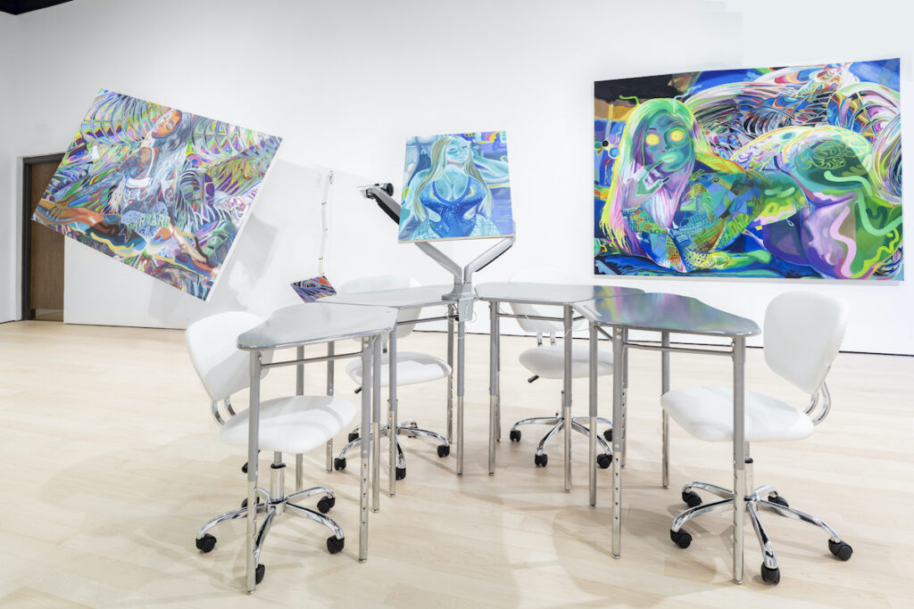 Four silver desks with white rolling chairs form a U-shape in the middle of a gallery. Three psychedelic-like canvases line the walls of the gallery, all abstract and multicolored.