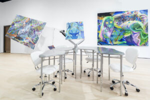 Four silver desks with white rolling chairs form a U-shape in the middle of a gallery. Three psychedelic-like canvases line the walls of the gallery, all abstract and multicolored.