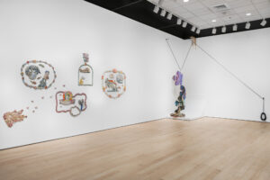 Installation view of ceramic works on a white wall, all including shapes or figures enclosed in a border. To the right is a sculpture of four stacked geometric flower-like shapes.